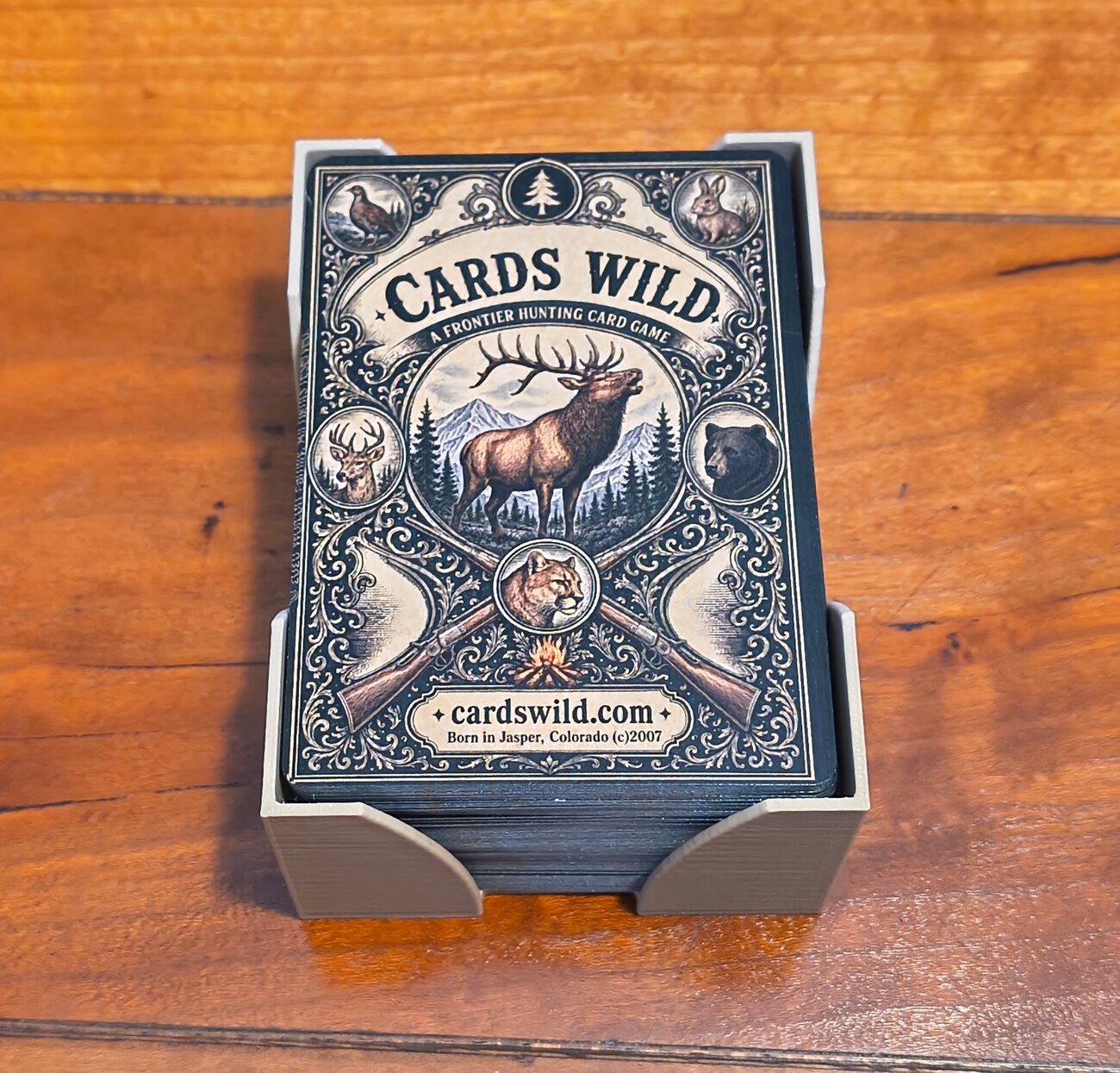 A Cards Wild deck standing in its 3D printed card holder on a wood table, the box front showing the elk engraving and the line Born in Jasper, Colorado 2007